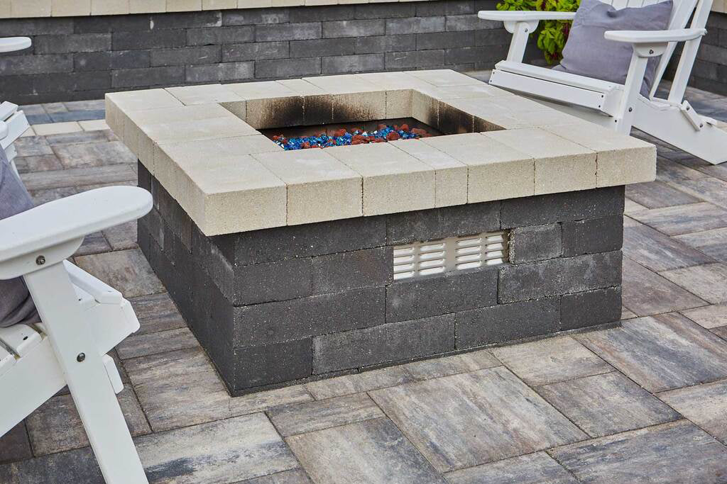 Summit Square Fire Pit on a patio in timeless with a retaining wall and chairs.
